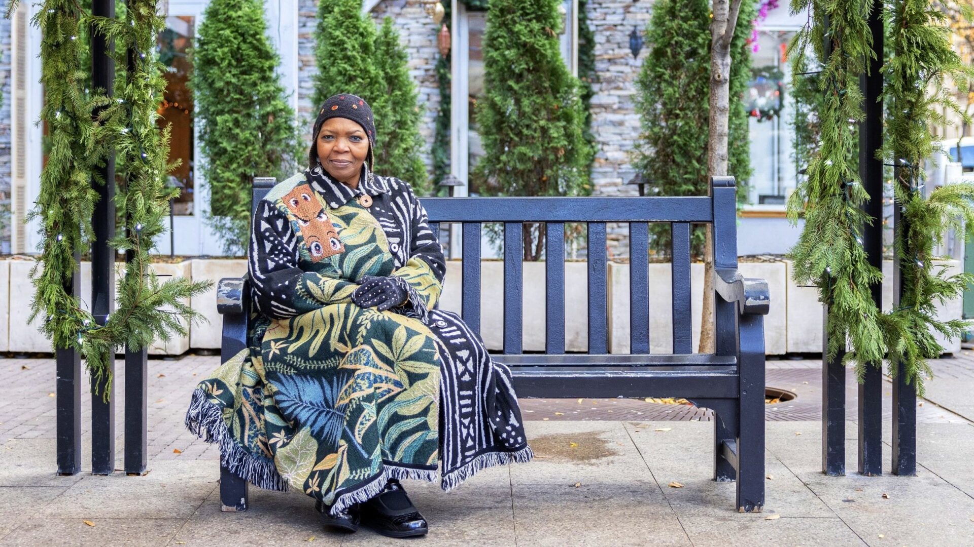 Sandy Baker seated on a bench between pine garlands in Hell's Kitchen, wearing a botanical-print coat