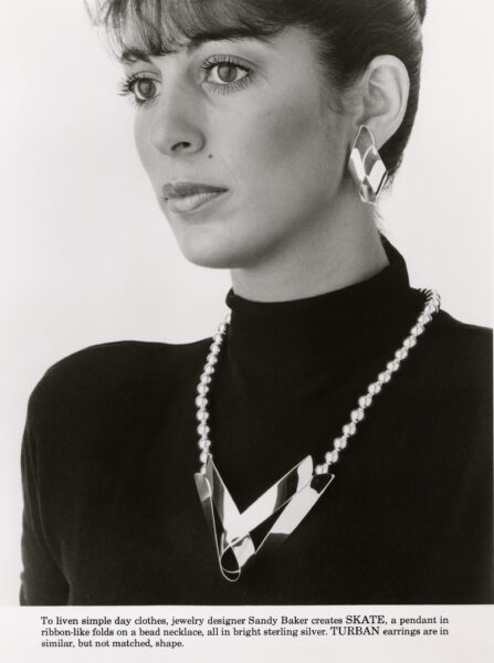 Her SKATE pendant on a ribbon-like fold, worn with TURBAN Earrings in a matching shape.